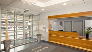 Aula, jossa on puinen vastaanottotiski, lasiovet ja vastaanottoalueelle sijoitettu matto, jossa lukee sana ”Vastaanotto”.