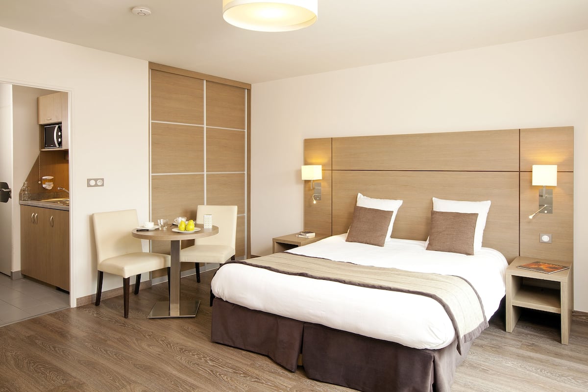 A hotel room with a bed, bedside tables, a desk with a chair, a small table with a chair, and a microwave in the kitchen area.