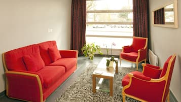 Een zithoek met een rode bank, een rode fauteuil en een klein tafeltje. Er is een raam met gordijnen, een plant op het tafeltje en een uitzicht op de straat.