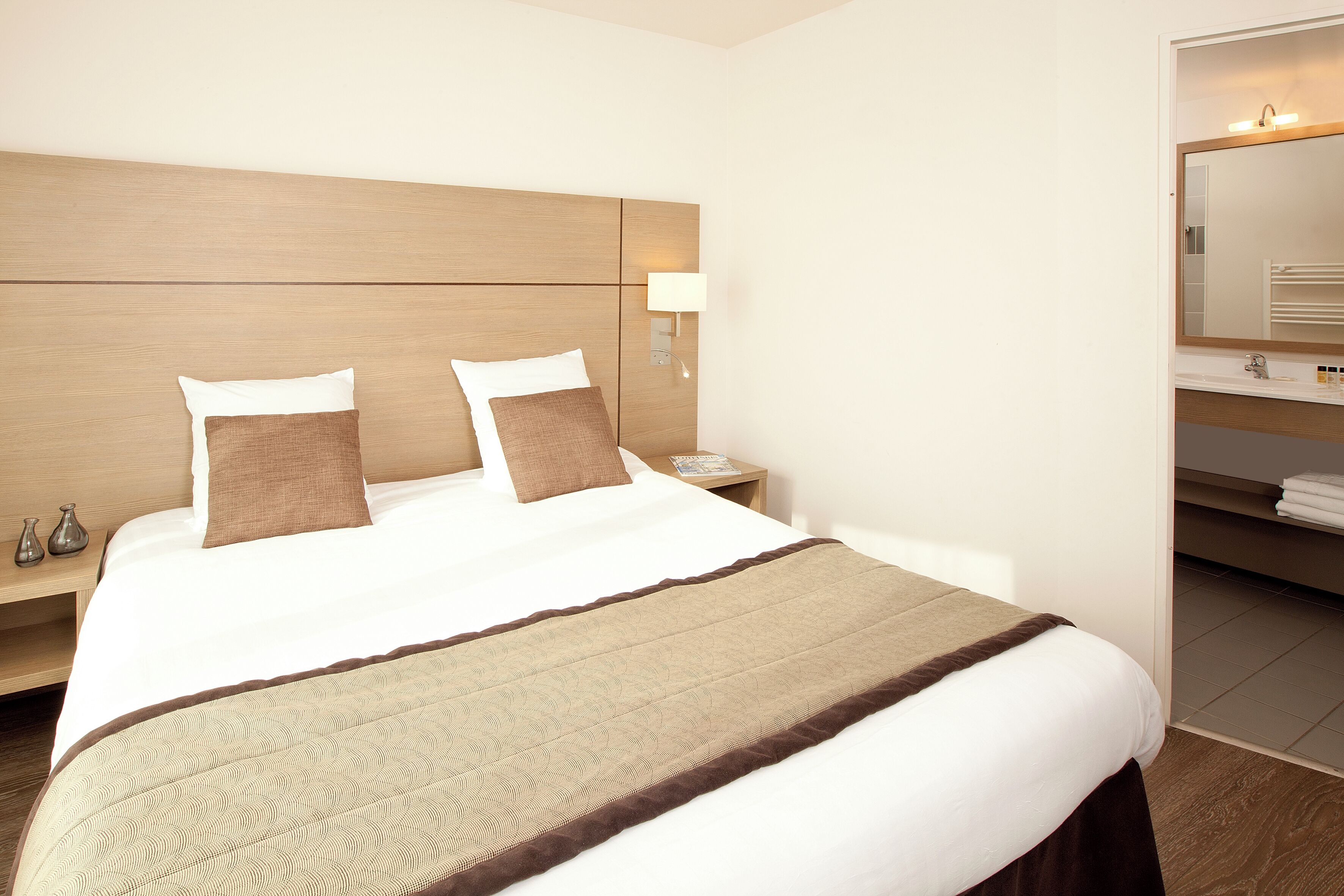 Ein Hotelzimmer mit einem großen Bett, einem Nachttisch mit Lampe und einem durchgegangene Tür sichtbaren Badezimmer.