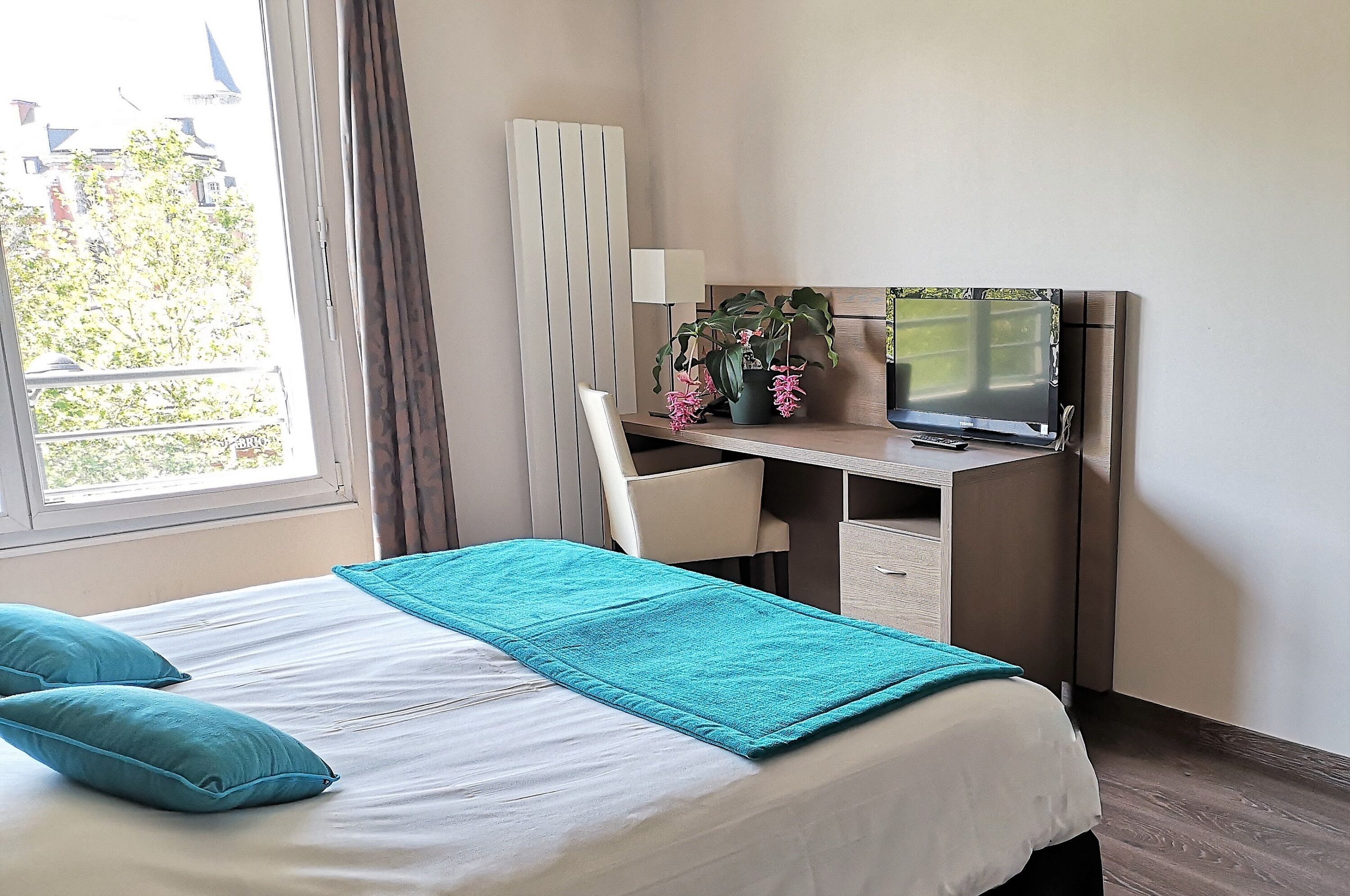 Ein Hotelzimmer mit Bett, Schreibtisch, Fernseher und Fenster mit Blick auf Gebäude und Bäume.