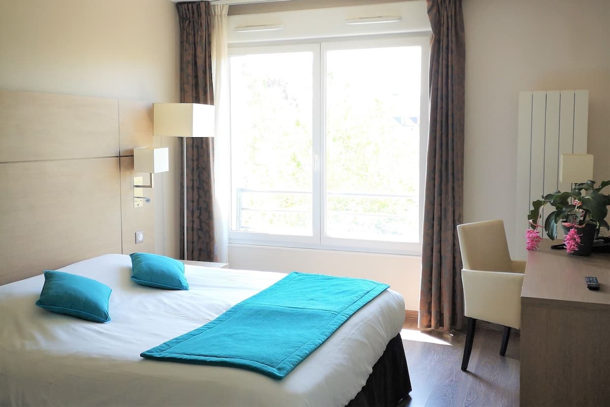 A hotel room with a bed, a desk, a chair, a lamp, and a window with curtains.
