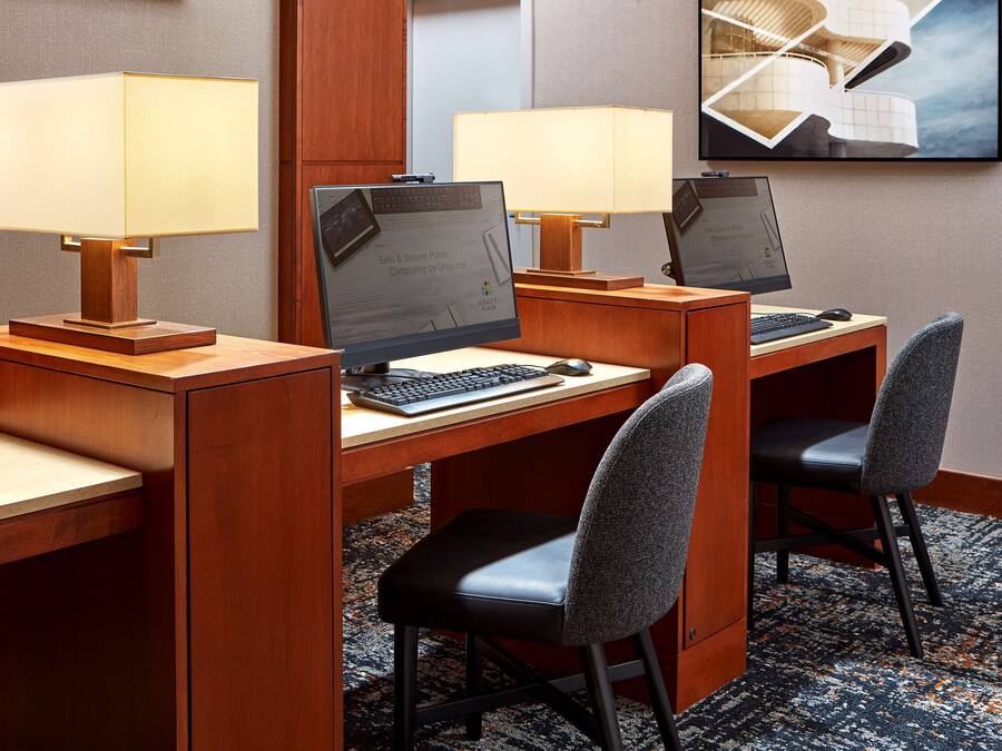 A hotel room with two desks, each with a computer, keyboard, and mouse. There are two chairs, one at each desk. The room has a carpeted floor and a framed picture on the wall.