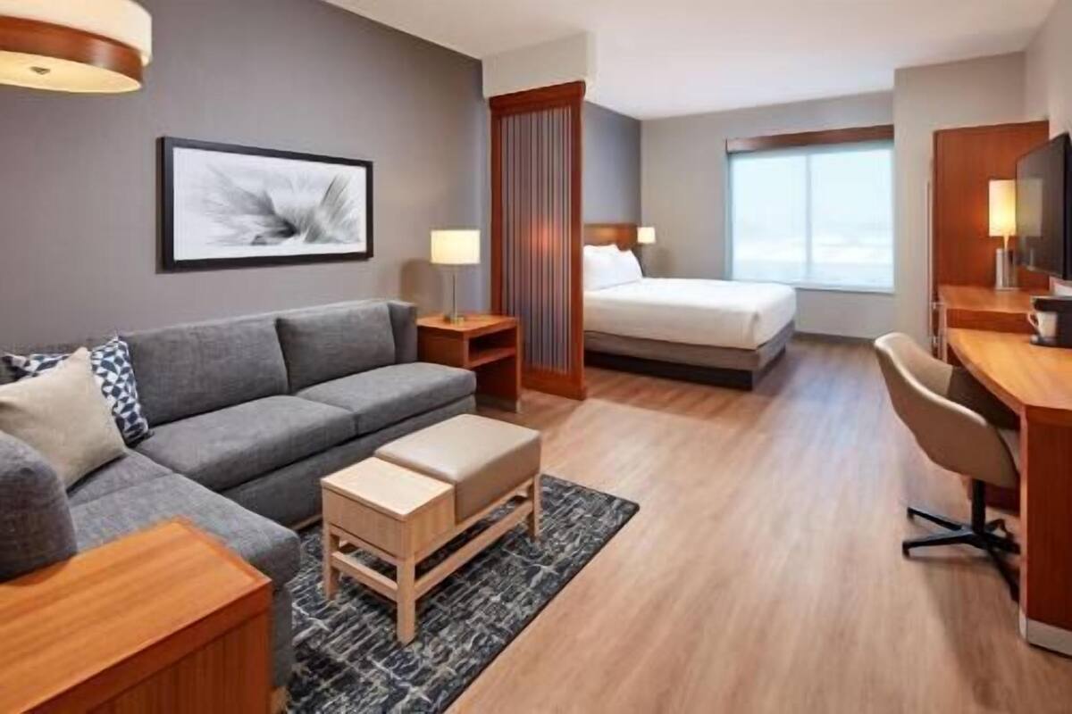 A hotel room with a grey sofa, a wooden desk, a bed, a TV, and a window.