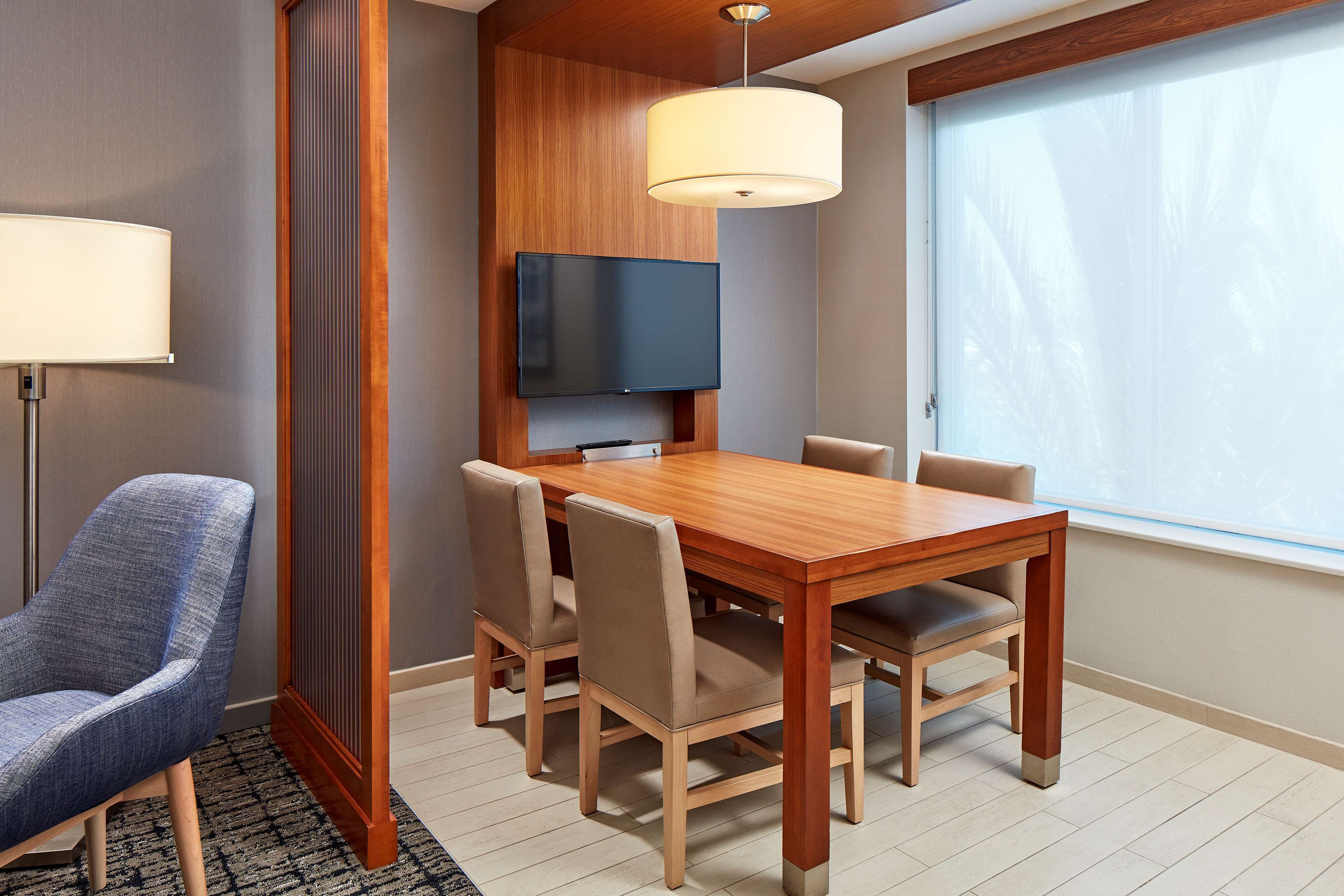 A hotel room with a wooden dining table, beige chairs, a flat-screen TV mounted on the wall, a large window with a view of palm trees, and a floor lamp.