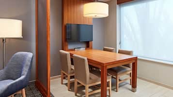 A hotel room with a wooden dining table, beige chairs, a flat-screen TV mounted on the wall, a large window with a view of palm trees, and a floor lamp.