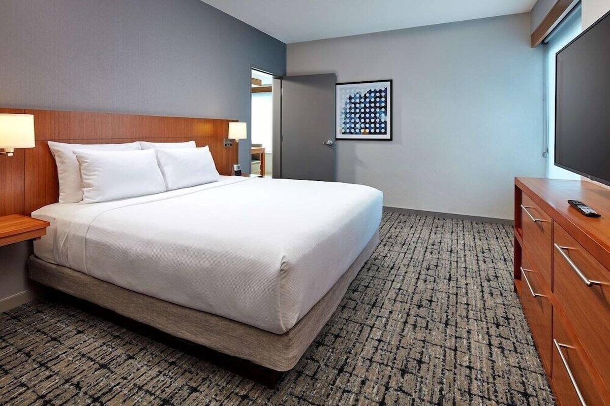 A hotel room with a bed, a wooden nightstand, a dresser, a flat-screen TV, and a framed picture on the wall.