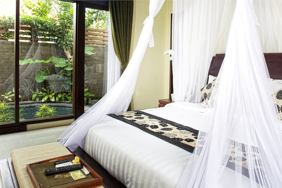 A bedroom with a bed under a canopy, a bedside table with a tray of items, and a view of greenery through a window.