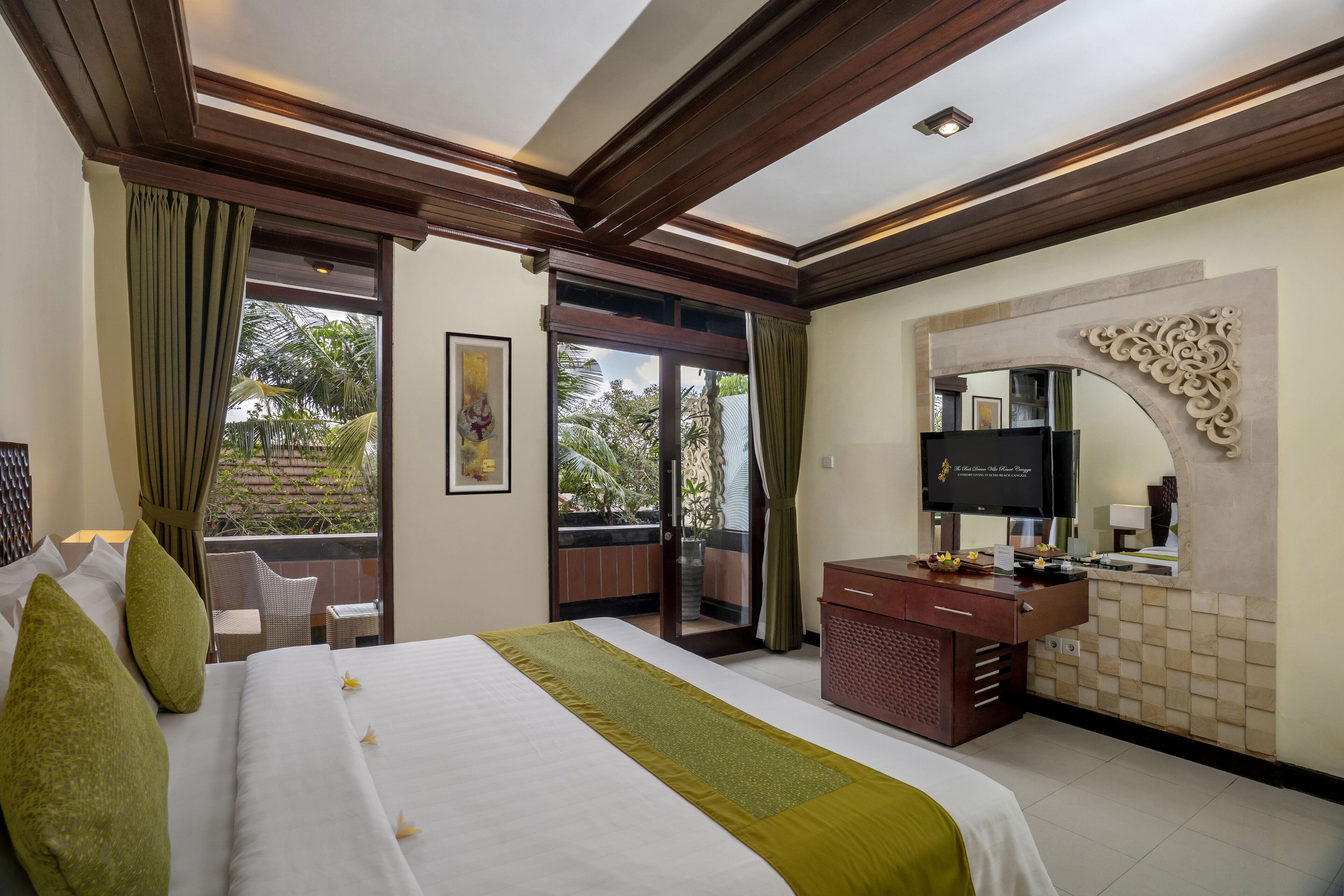 A hotel room with a large bed, a wooden ceiling, a balcony with a view of palm trees, a TV, and a bathroom with a mirror and sink.