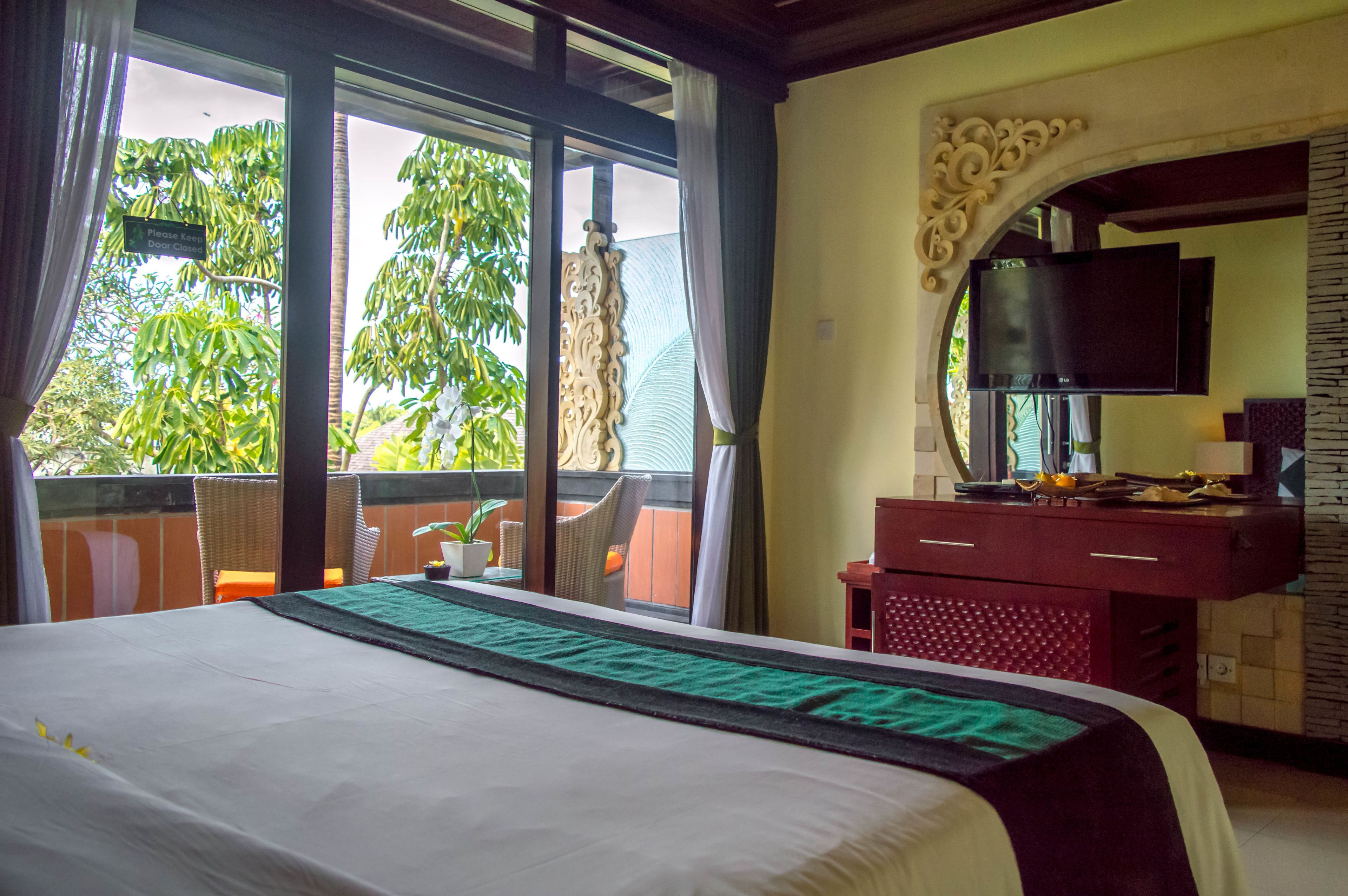 Habitación de hotel con una cama grande, un televisor en la pared, un balcón con sillas de mimbre y vistas a árboles.