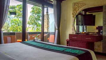 Habitación de hotel con una cama grande, un televisor en la pared, un balcón con sillas de mimbre y vistas a árboles.