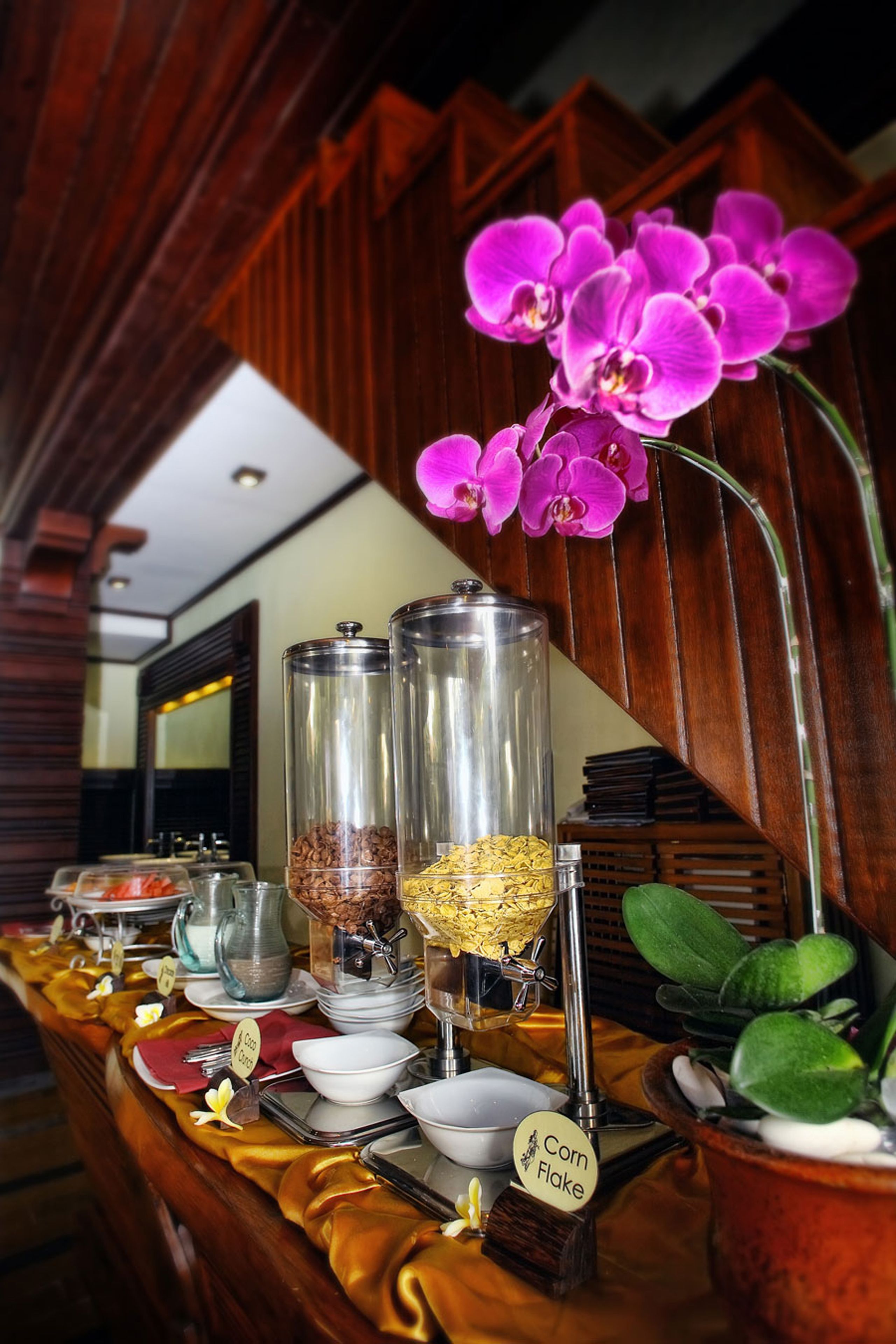 A buffet setup with various food containers, a sign indicating 'Corn Flake', and a potted plant.