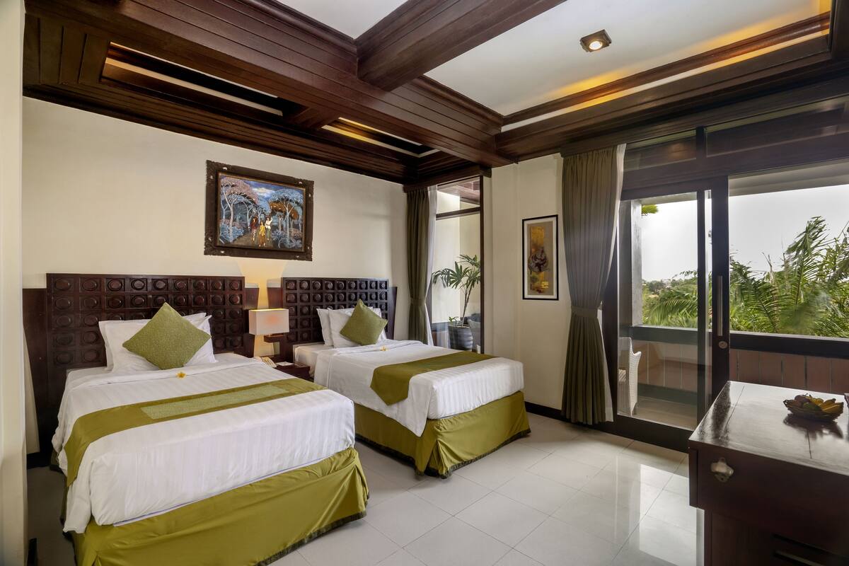 A hotel room with two beds, wooden headboards, a large window with a view, and a desk with a bowl of fruit.