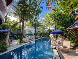 A pool area with lounge chairs and umbrellas, surrounded by lush greenery and a clear blue sky.