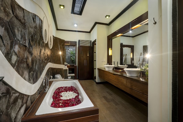 A modern bathroom with a stone wall, a large bathtub filled with rose petals, and a wooden vanity with a mirror.