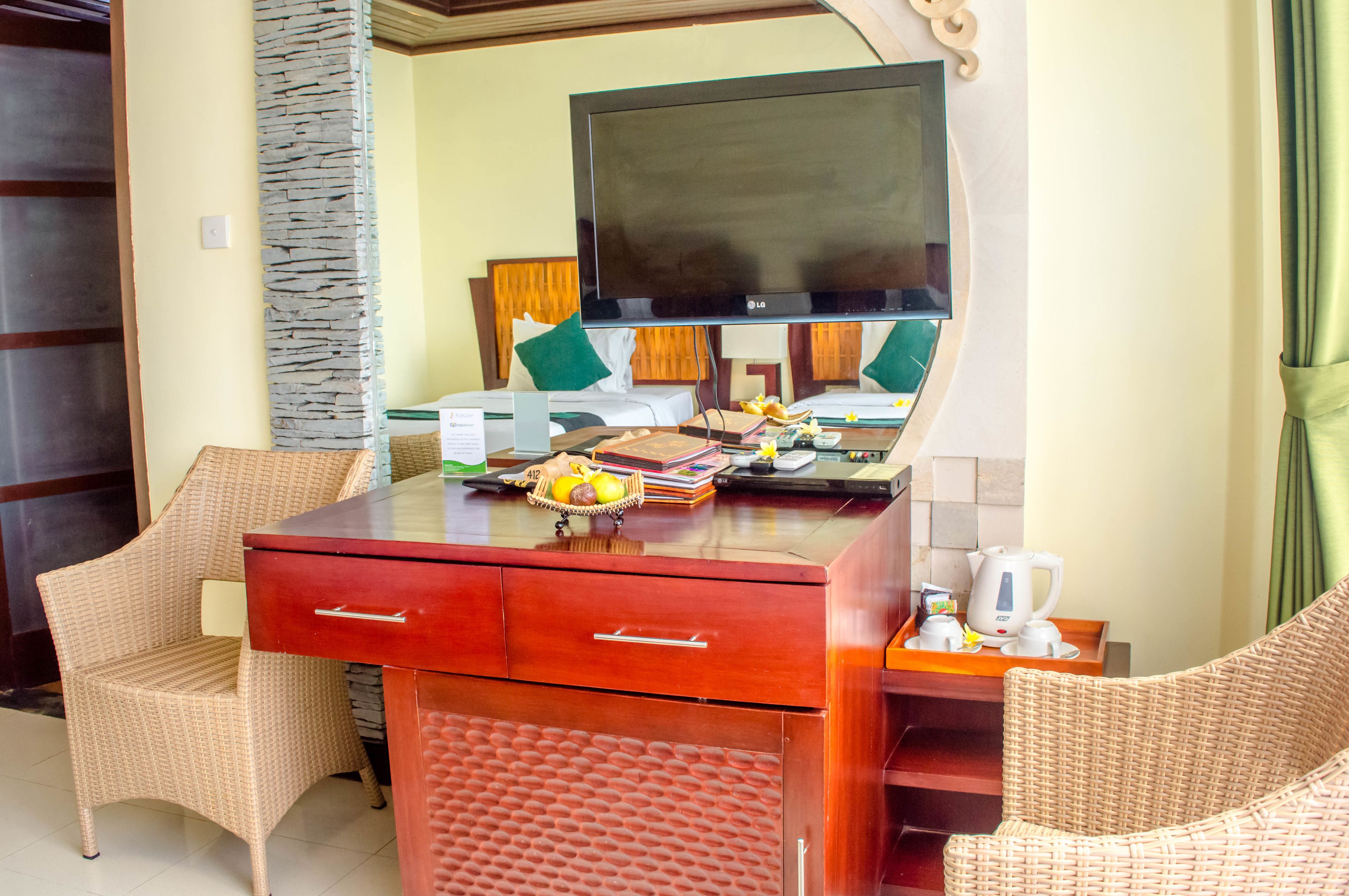 Une chambre d’hôtel avec un bureau en bois, deux fauteuils en osier, une télévision et un lit avec des oreillers verts.