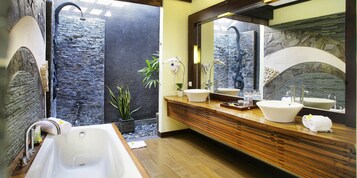 A modern bathroom with a large bathtub, two sinks, a shower, and a stone wall.