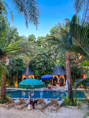A pool area with lounge chairs and umbrellas, surrounded by lush greenery and palm trees.