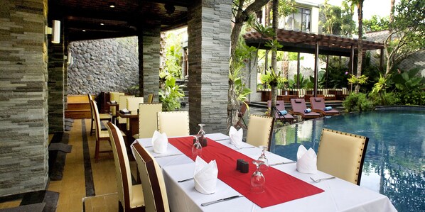 A restaurant with a pool view, featuring a stone wall, wooden furniture, and a red table runner.