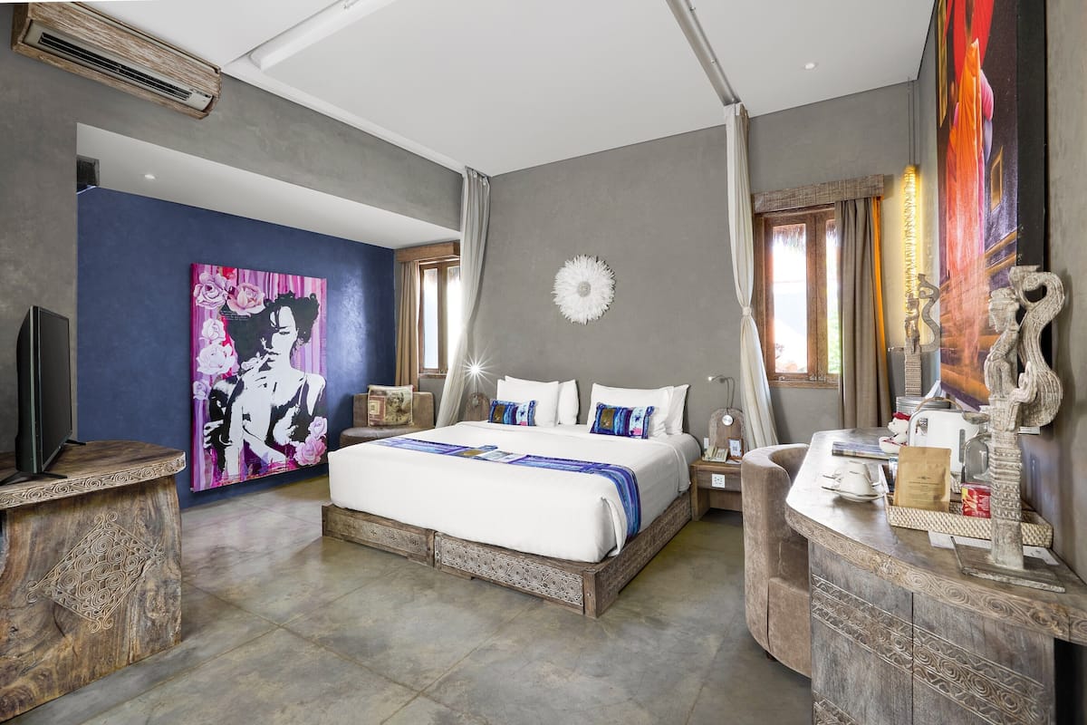 A bedroom with a bed, a TV, a painting, a clock, and a desk with a lamp.