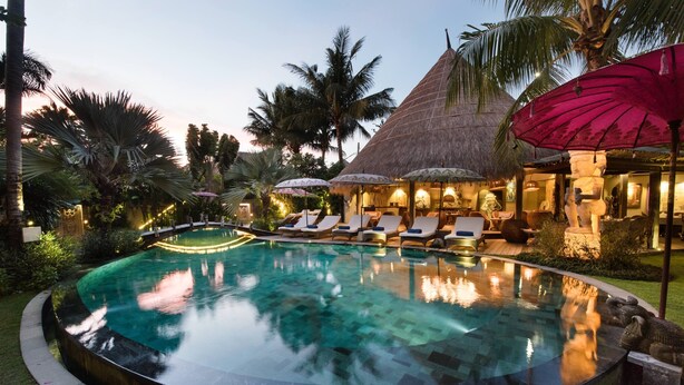 A swimming pool with lounge chairs and umbrellas, surrounded by palm trees and a thatched-roof building. - Blue Karma Dijiwa Seminyak (Seminyak)