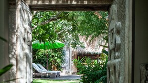 A pathway leading to a pool area with a thatched roof structure, surrounded by lush greenery and a stone wall. - Blue Karma Dijiwa Seminyak (Seminyak)