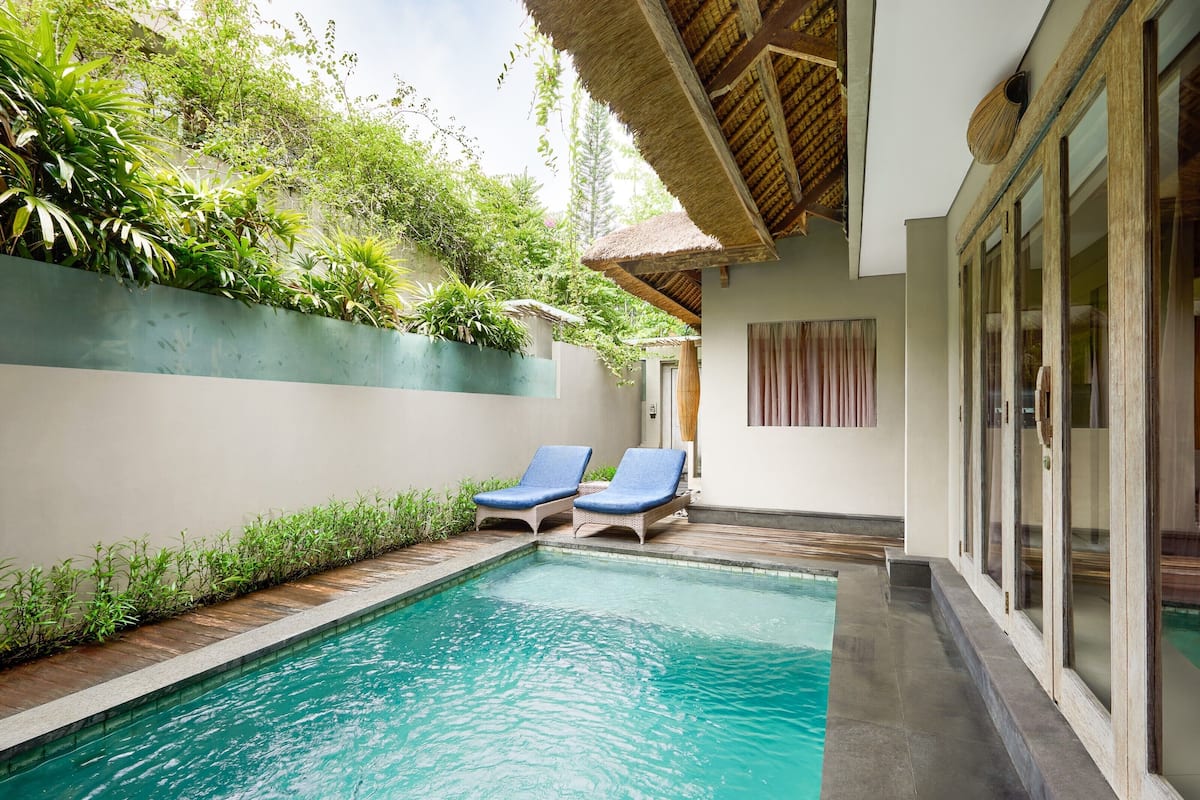 A pool area with two lounge chairs, a thatched roof, and lush greenery.