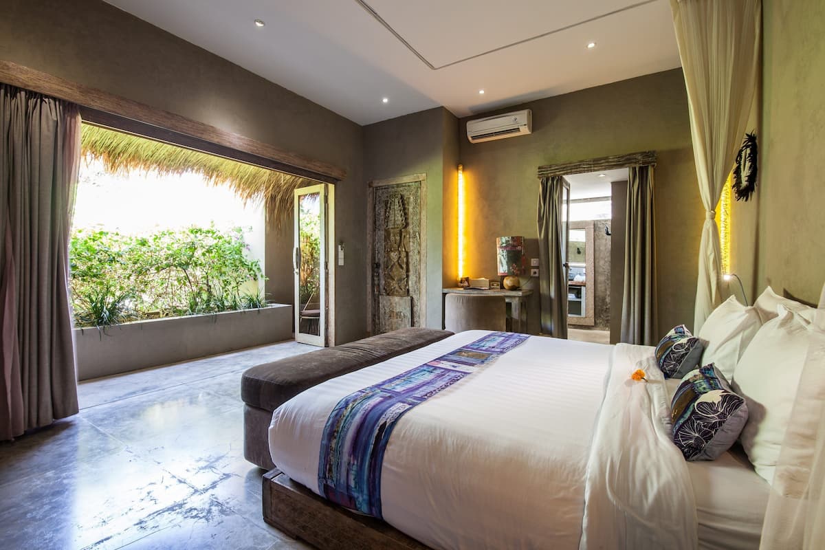 A bedroom with a large bed, a nightstand, a mirror, and a view of greenery outside.