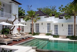 Uma área de piscina com espreguiçadeiras e guarda-sóis, cercada por palmeiras e um edifício branco com janelas amplas.