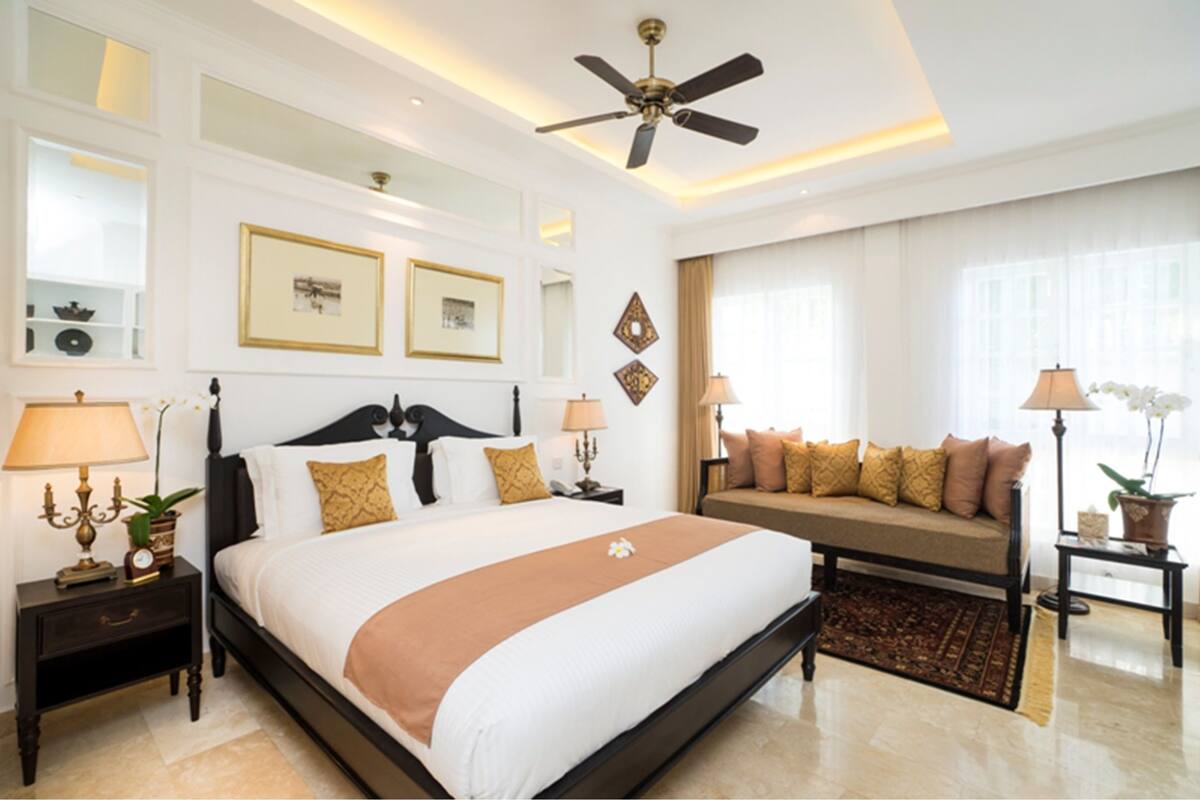 Une chambre d’hôtel avec un grand lit, un canapé, deux tables de chevet avec des lampes, un ventilateur de plafond, des tableaux encadrés au mur et une fenêtre avec des rideaux.