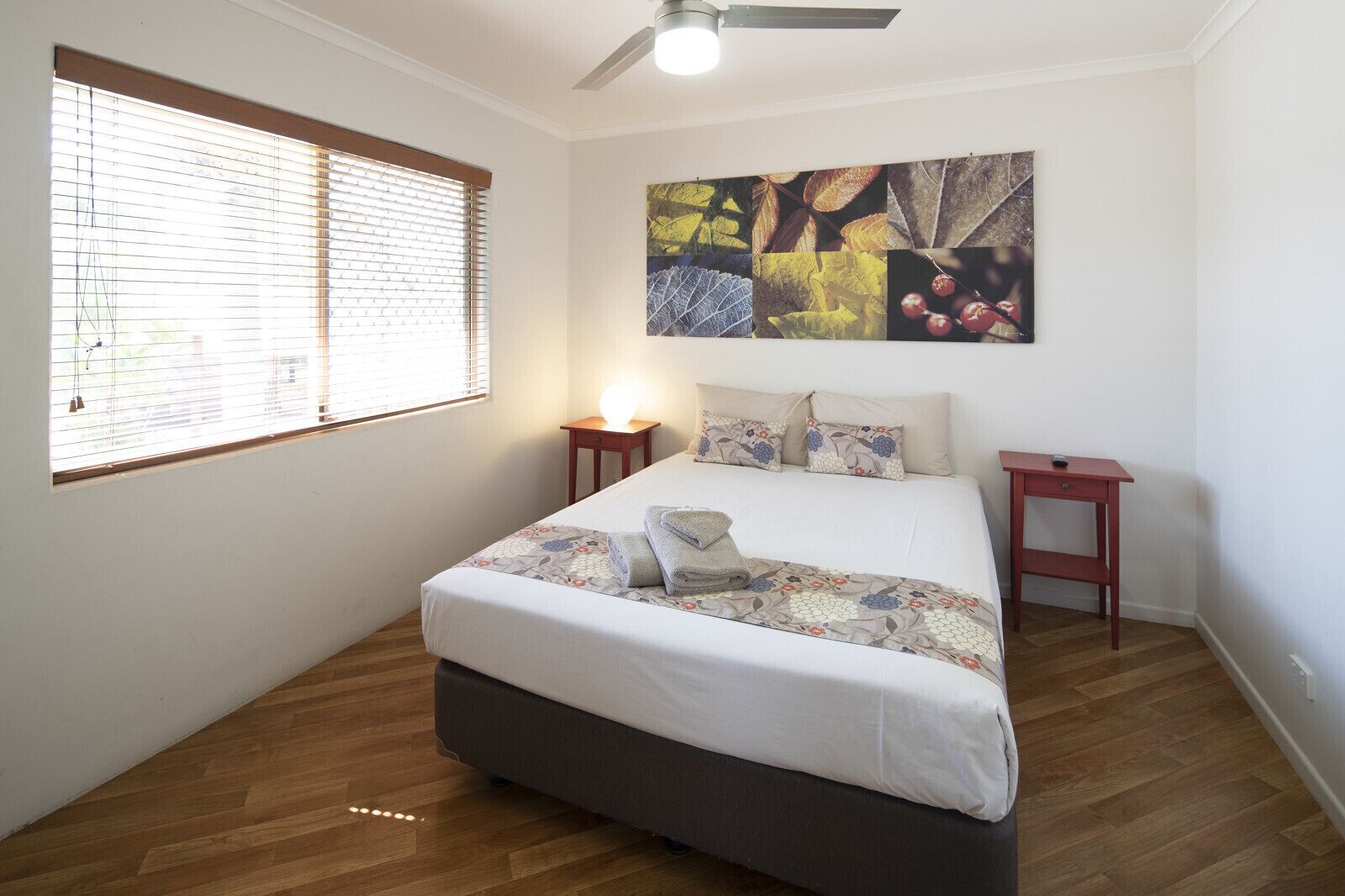 Un dormitorio con cama, mesita de noche, ventilador de techo y ventana con persianas.