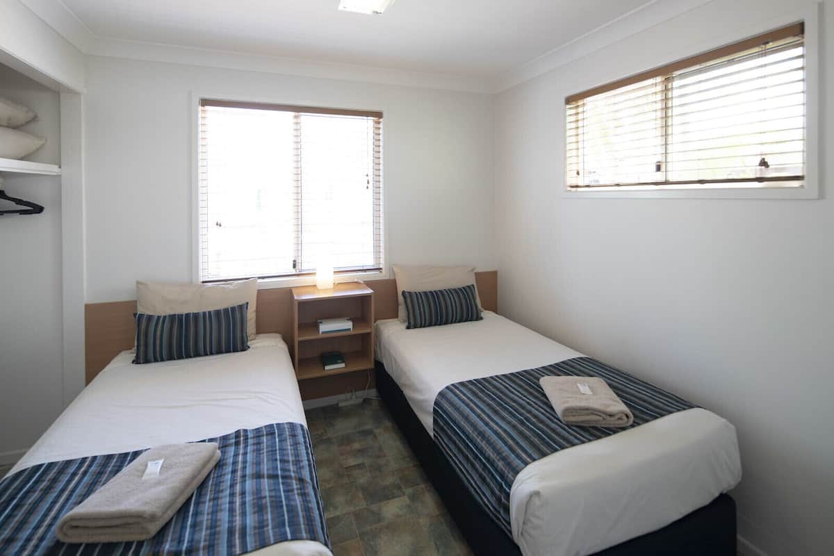 Une chambre avec deux lits simples, une petite étagère en bois et une fenêtre avec des stores.