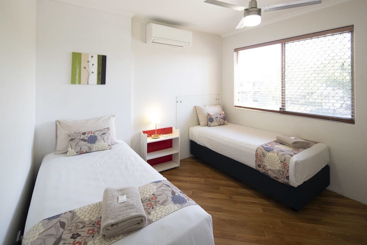 Une chambre avec deux lits, un ventilateur de plafond, un appareil de climatisation, une table de chevet avec une lampe et une fenêtre avec des stores.