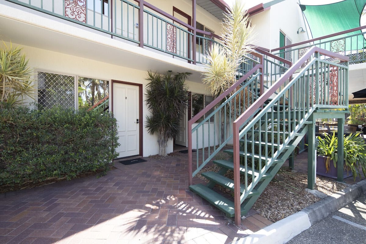 Un immeuble à plusieurs étages, doté d’un escalier, d’une porte et entouré de végétation.
