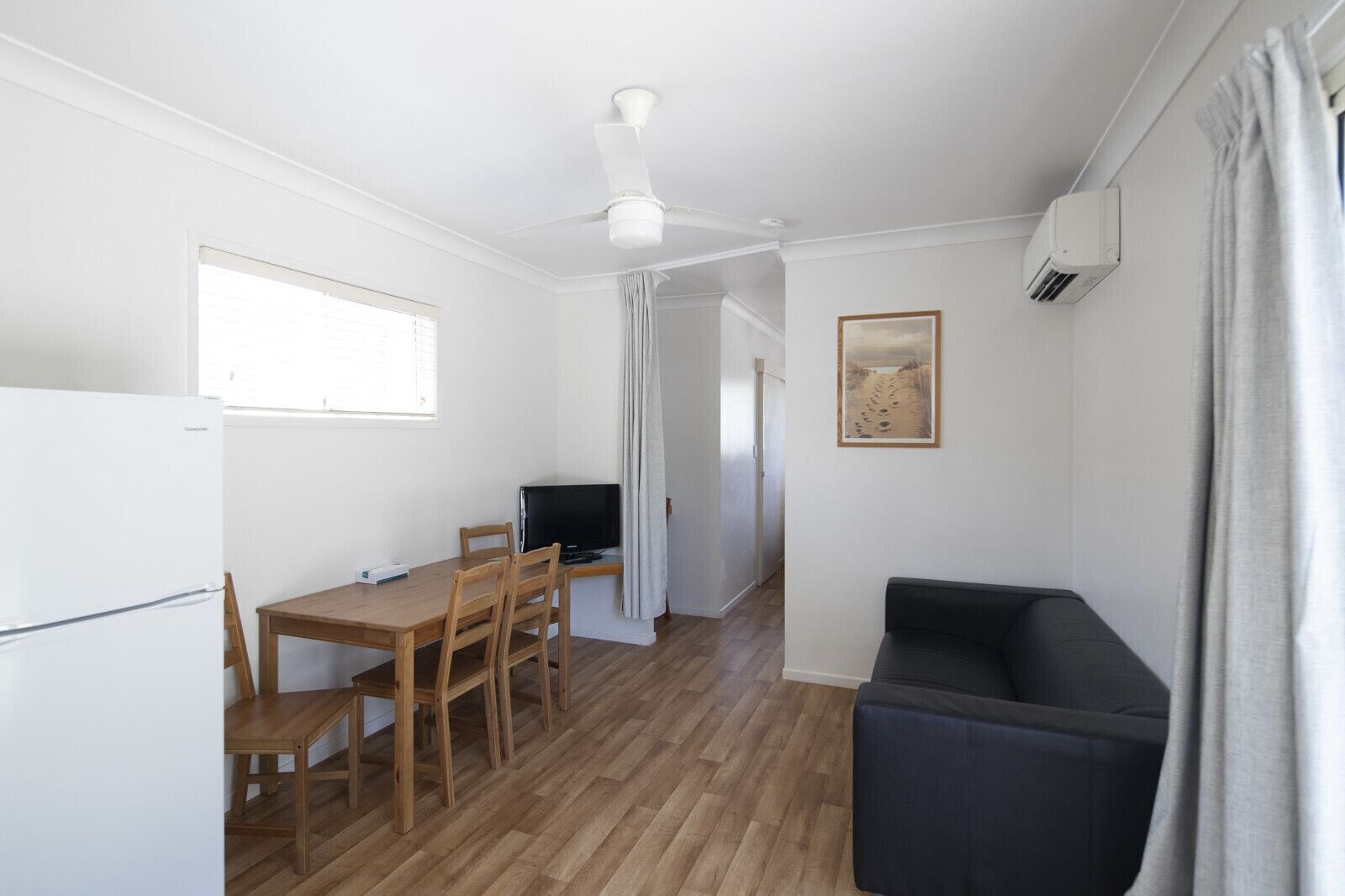 Una habitación compacta con una mesa de comedor de madera, un sofá negro, un ventilador de techo, un televisor y un cuadro en la pared.