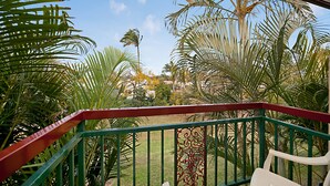 Un balcon doté d’une balustrade verte et rouge, donnant sur un jardin planté de palmiers et surplombant un ciel d’un bleu limpide.