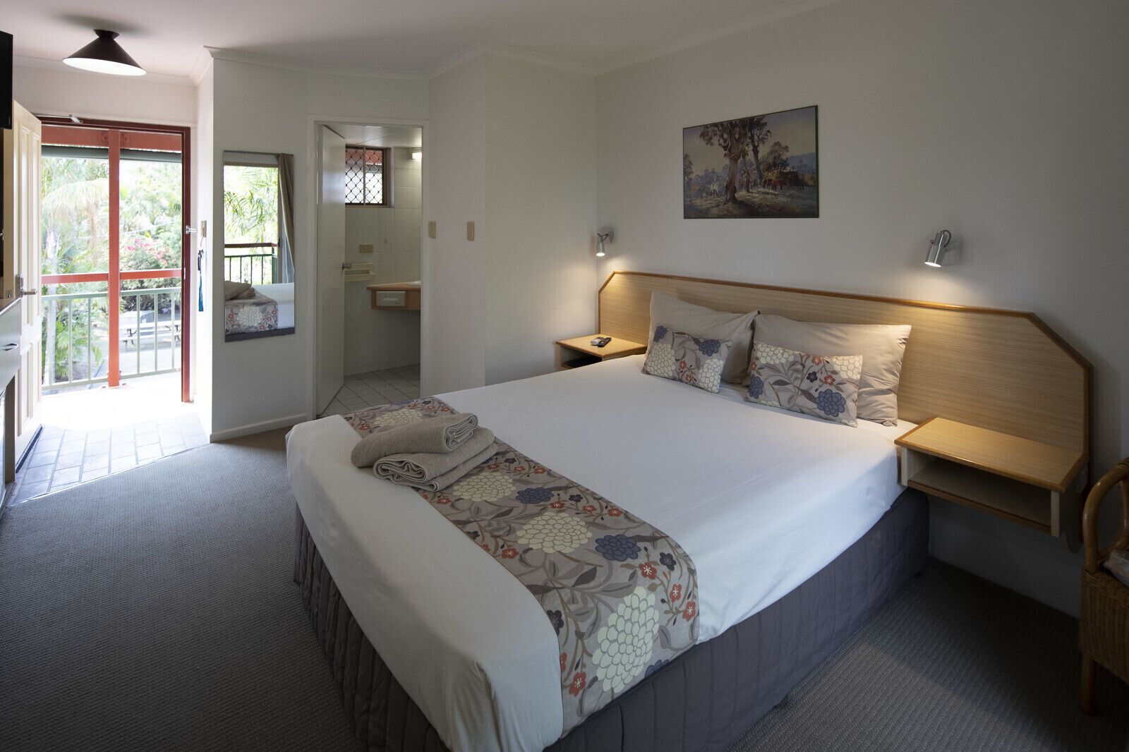 A spacious bedroom with a large bed, a nightstand, a wall-mounted lamp, and a painting.