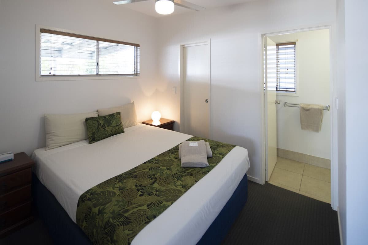 Une chambre à coucher comprenant un lit, une table de chevet, une fenêtre et une porte donnant sur une salle de bain.