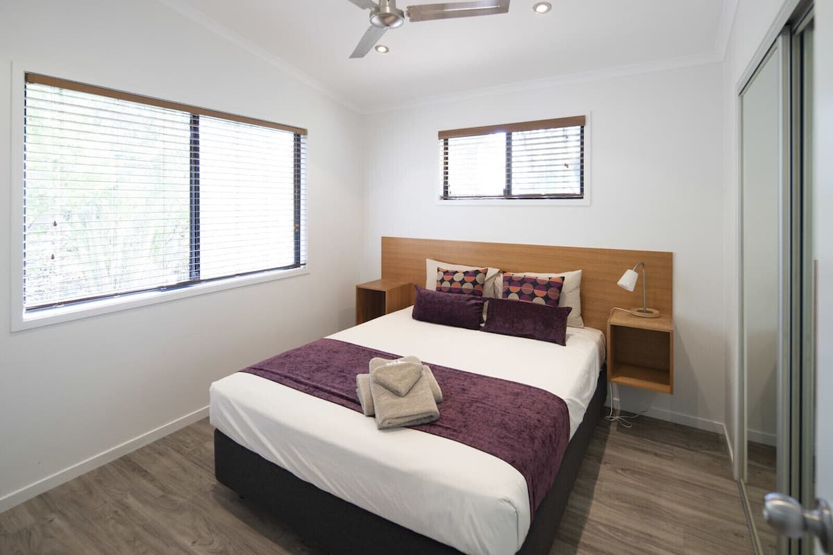 Une chambre moderne avec un grand lit, deux tables de chevet, un ventilateur de plafond et deux fenêtres équipées de stores.