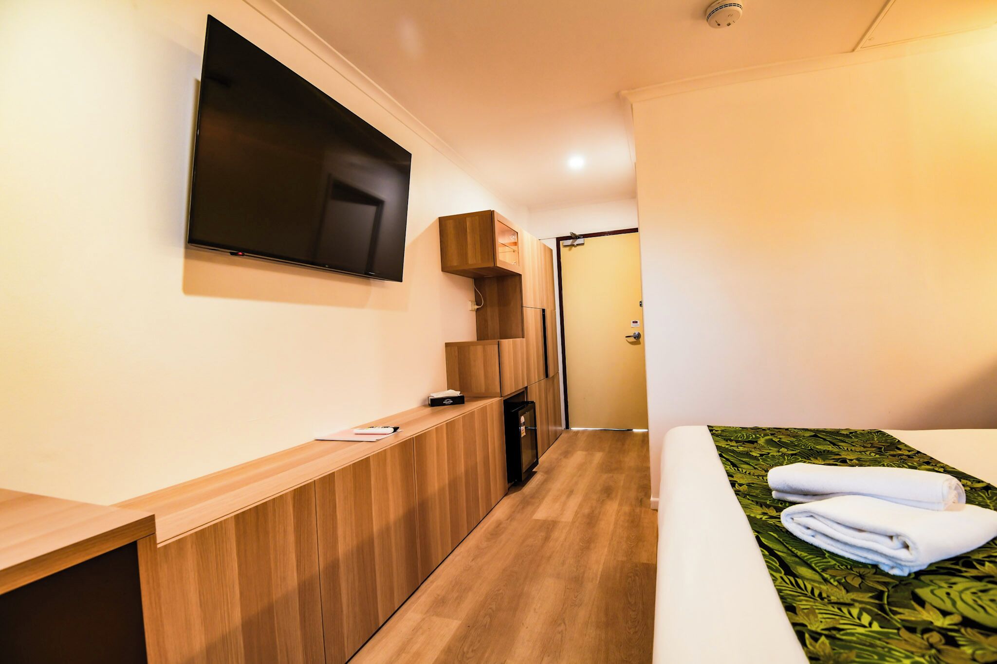 Une chambre d’hôtel avec un bureau en bois, un téléviseur à écran plat fixé au mur, un lit recouvert d’une courtepointe à motifs de feuilles vertes, et une porte visible au fond.