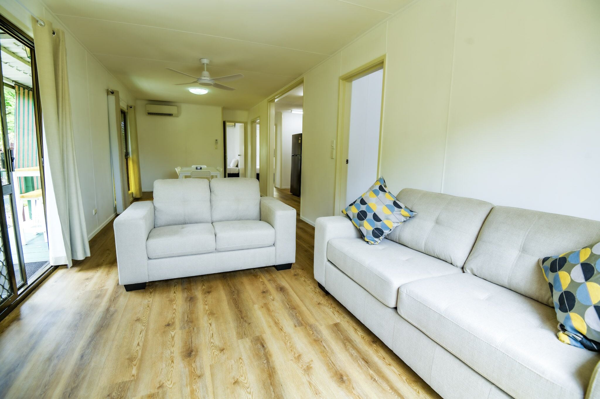 A spacious living area with a white sofa and a matching armchair, a ceiling fan, and a wooden floor.