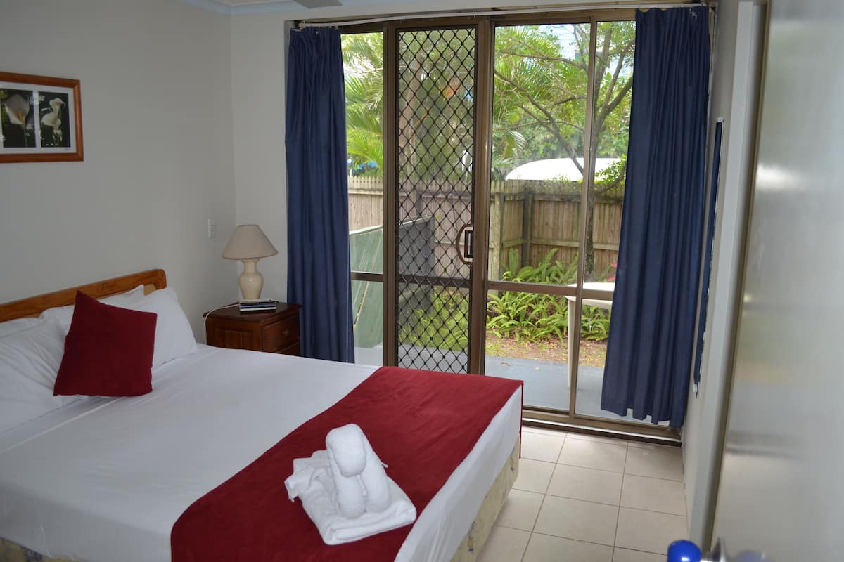 Habitación de hotel con cama, mesita de noche, lámpara y vistas a un área exterior con vegetación a través de una puerta corrediza de vidrio.