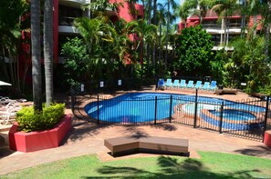A pool area with a blue pool, surrounded by palm trees and a fence. There are lounge chairs and a bench around the pool. - Enderley Gardens Resort (Surfers Paradise)