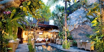 Uma área de refeições ao ar livre com mobiliário de vime, piscina e plantas tropicais.