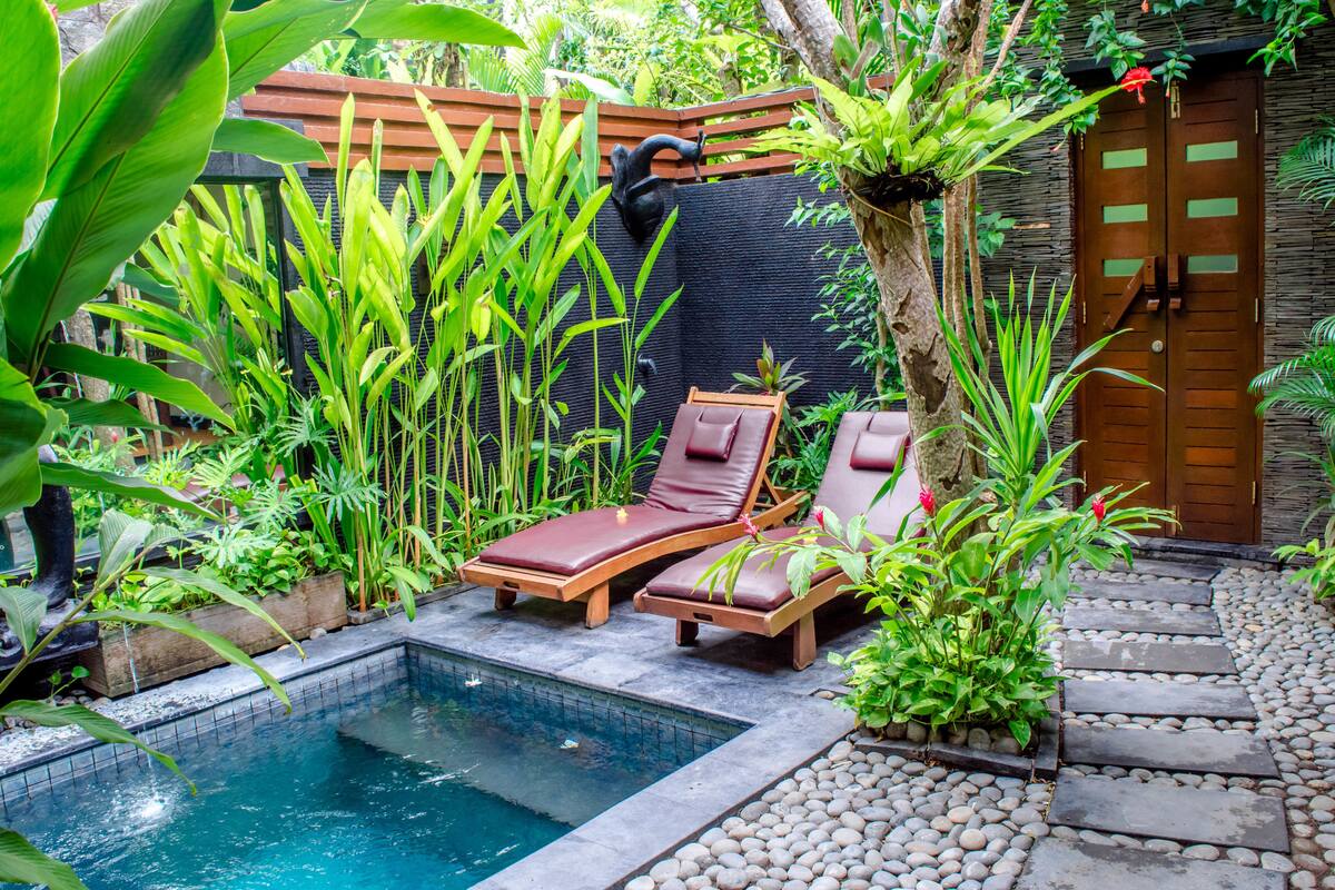A private pool area with two wooden lounge chairs, surrounded by lush greenery and a stone pathway.