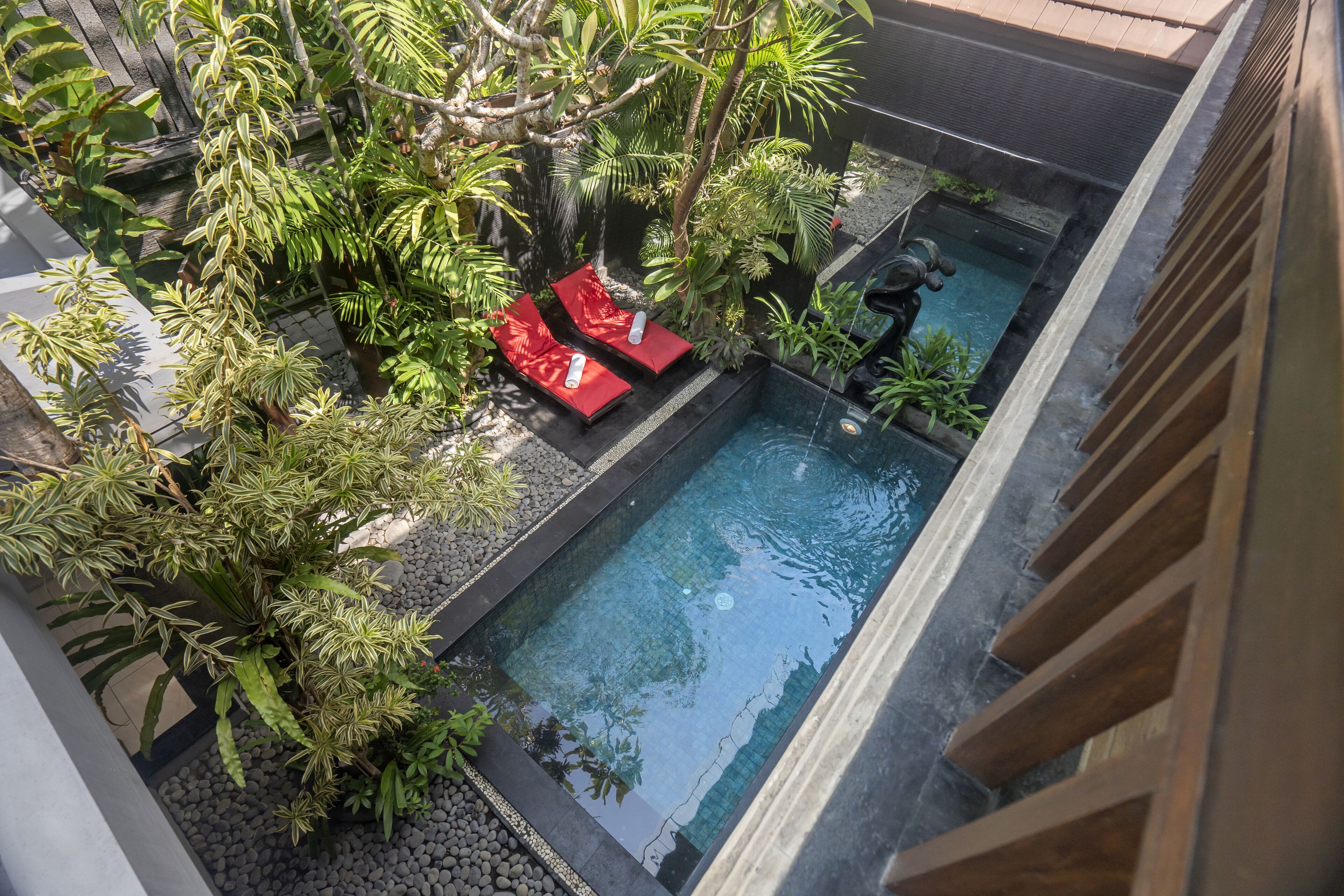 A rooftop pool with two red lounge chairs, surrounded by lush greenery and a small waterfall feature.