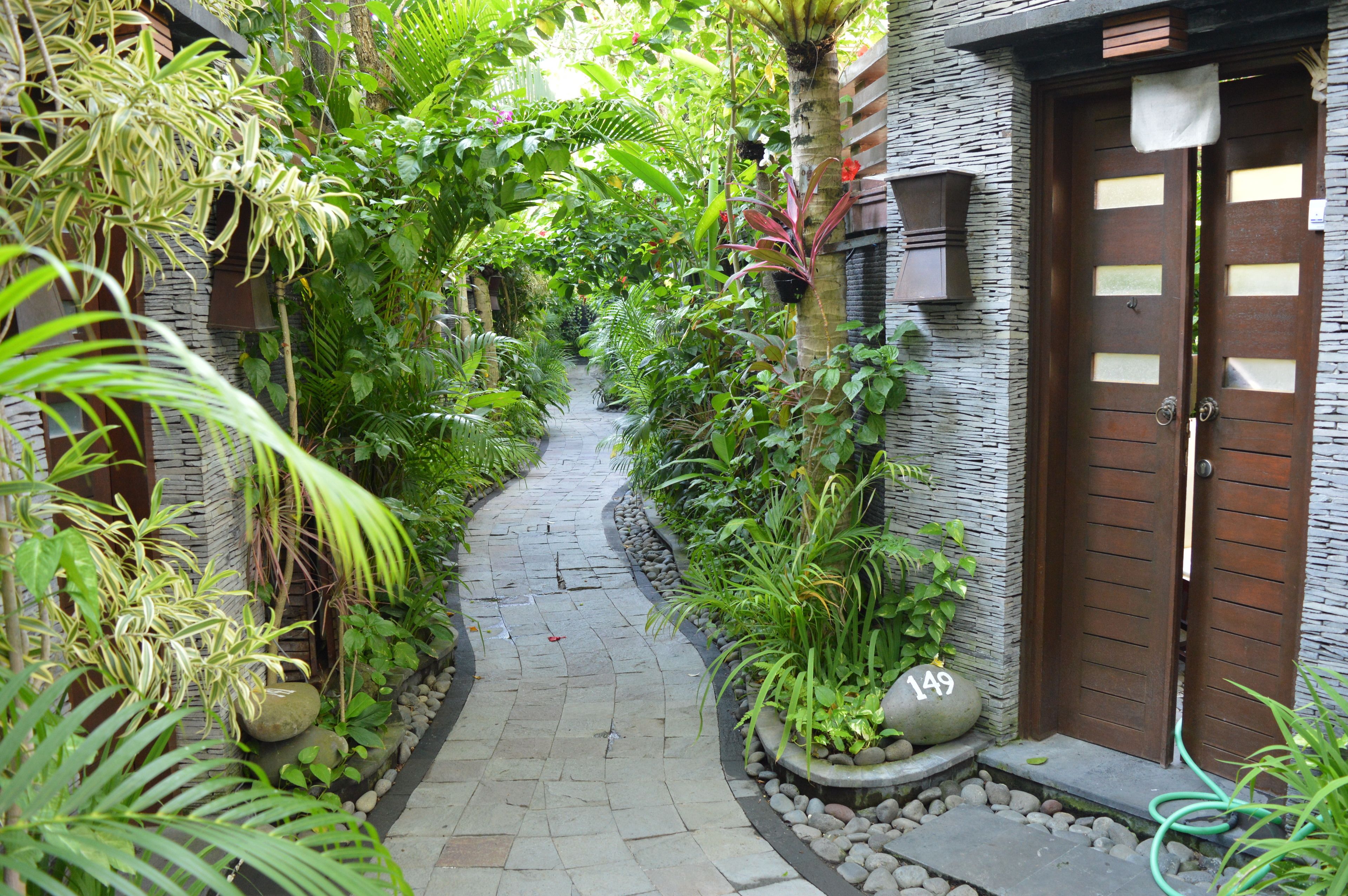 A narrow stone pathway flanked by lush greenery and a stone building with a wooden door.