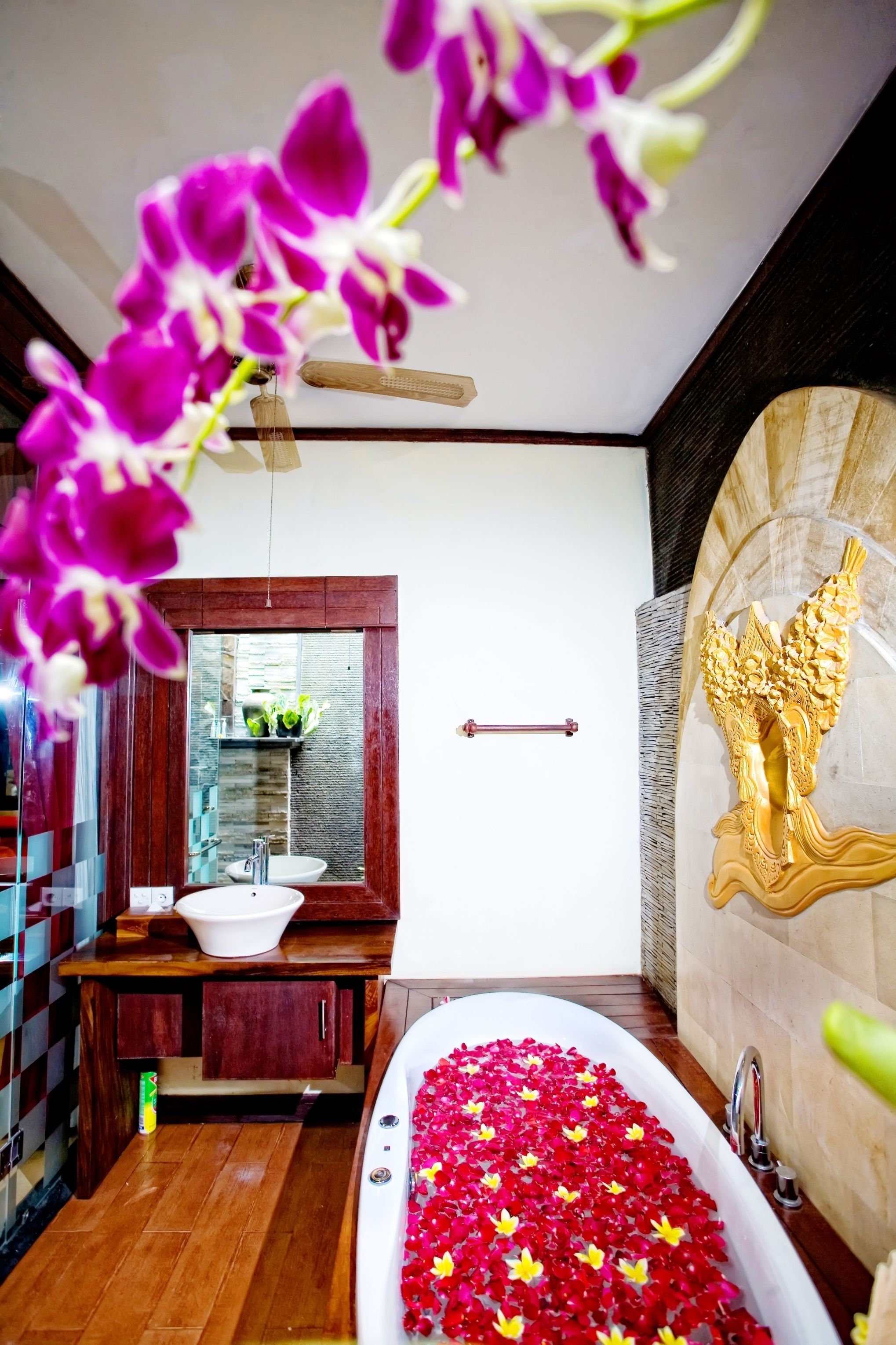 A bathroom with a bathtub filled with red and yellow flower petals, a white sink, and a wooden vanity.