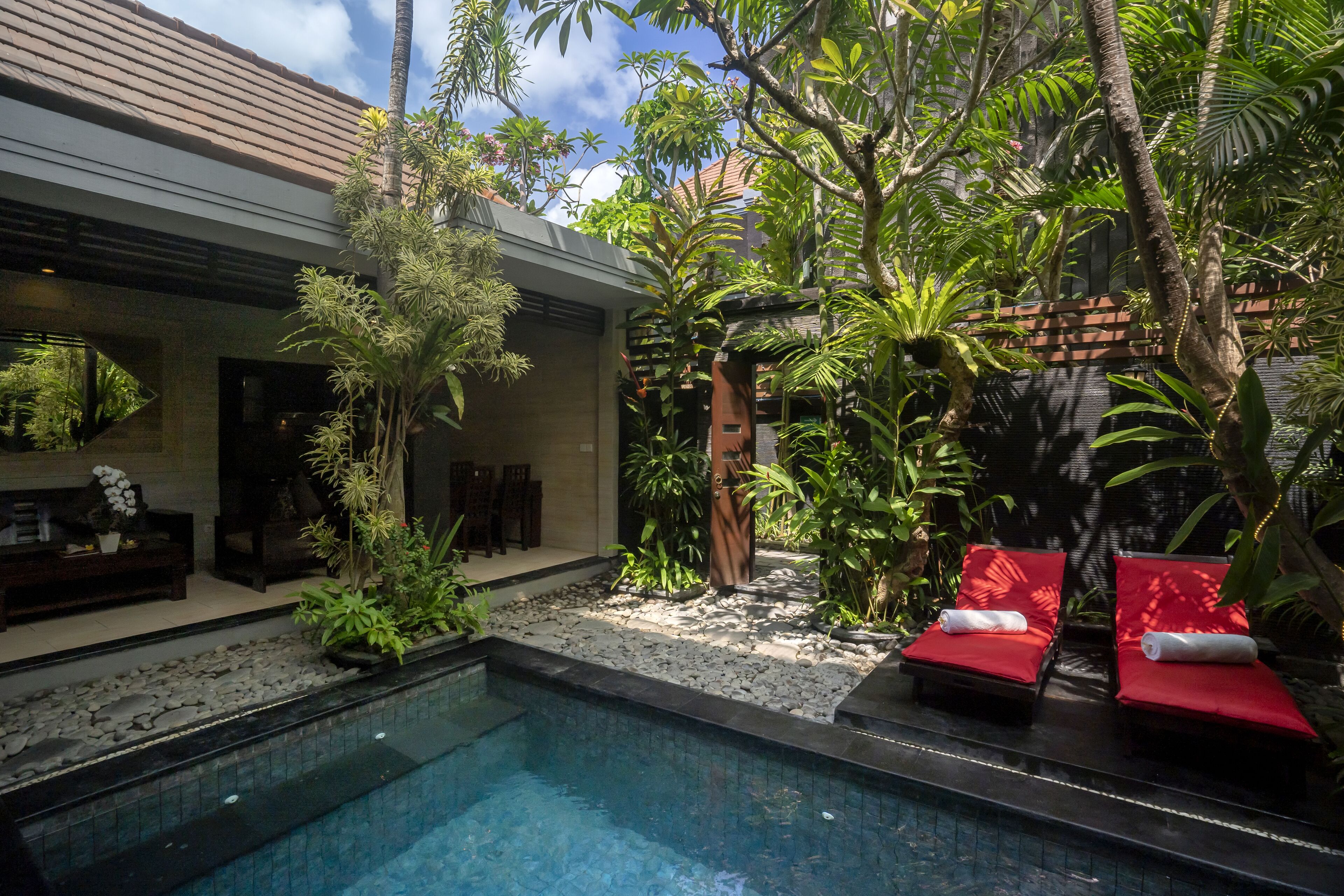 Un área de piscina con una piscina azul cristalina, rodeada de plantas tropicales y una tumbona roja.