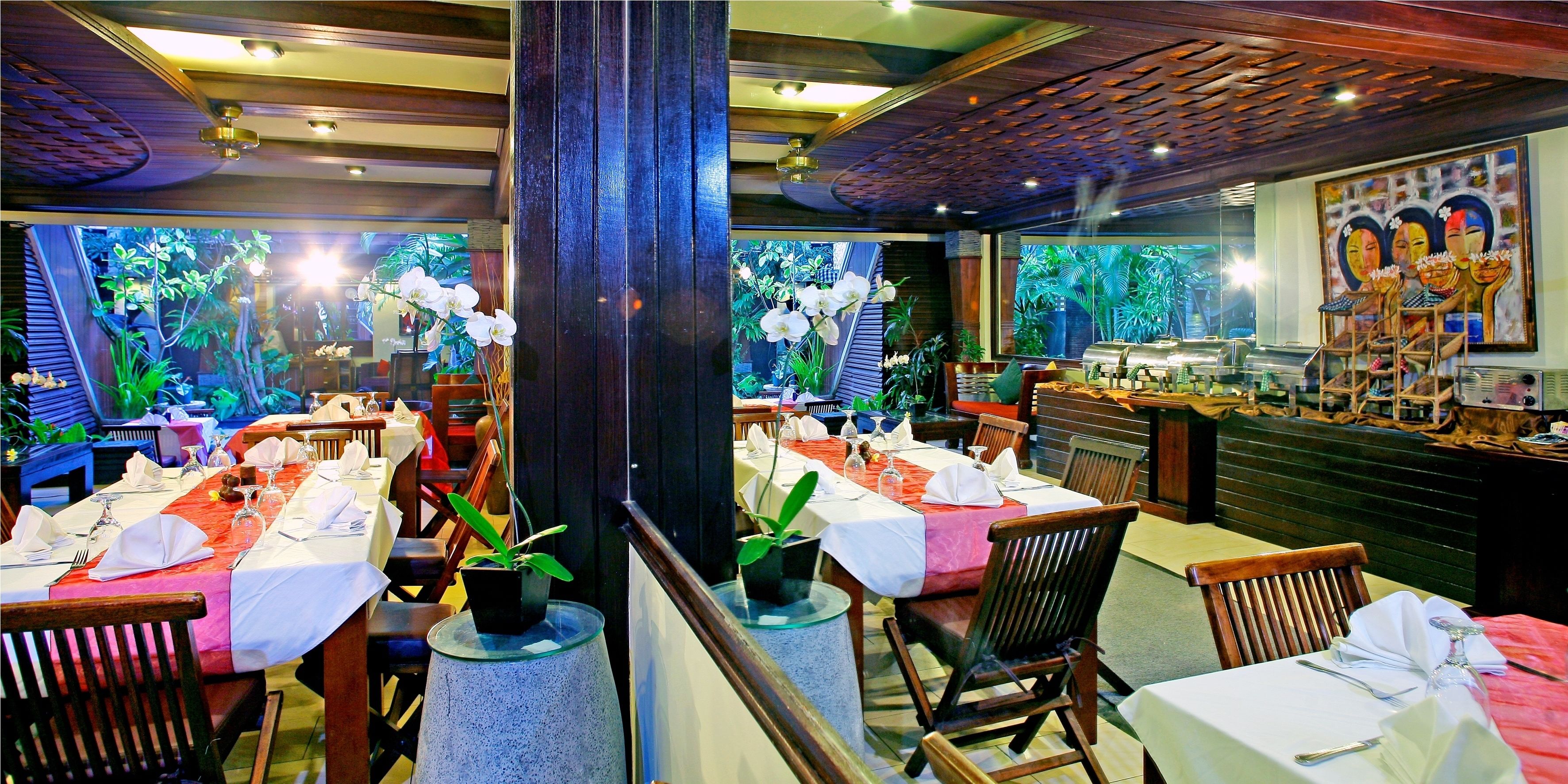 Un restaurant où les tables sont dressées pour le service, agrémenté de plantes en pot et doté d’une grande fenêtre offrant une vue sur un paysage tropical.